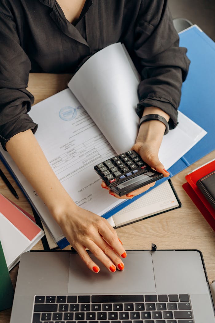 A business professional uses a calculator among documents and laptop on a desk for financial tasks.